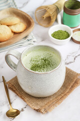 A mug of hot matcha green tea with mousse & its powder and spoon, chasen whisk and cookies in background on a table. It is rich in catechins, a class of plant compounds in tea as natural antioxidants
