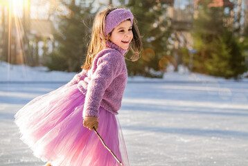 little girl in pink sweater and full skirt rides on sunny winter day on an outdoor ice rink in park