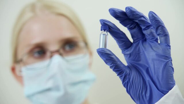 A female doctor in a medical mask and rubber gloves holds a small ampoule with a blue drug in her hands. Vaccination against dangerous diseases, strengthening immunity. Close-up, 4K UHD.