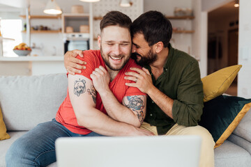 Young happy married gay couple sitting in their living room and having a video call with loved ones. Even though it's a coronavirus pandemic, there is always a way to spread happiness loved ones.