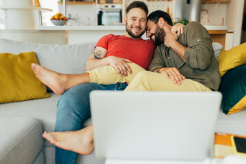 Happy married gay couple in love sitting in their cozy living room and watching movies on the laptop. They are cuddling.