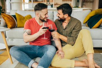 Overjoyed married gay couple in love sitting at home on the floor and toasting with red wine. They...