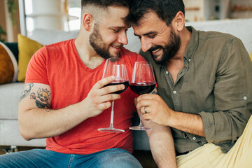 Overjoyed married gay couple in love sitting at home on the floor and toasting with red wine. They are celebrating their anniversary. - obrazy, fototapety, plakaty