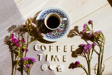 A cup of Turkish coffee on a white table with a silver tablecloth. Coffee time text inscription, smiley face, heart, home symbols top view. Dried purple lilac flowers flat lay. Beautiful composition.