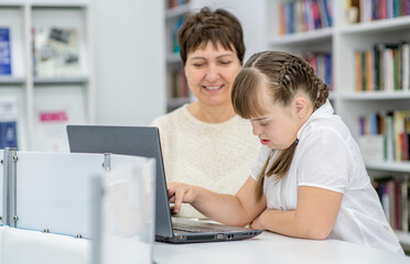 Smiling senior woman teaches a young girl with down syndrome to use a laptop at school. Education for disabled children concept