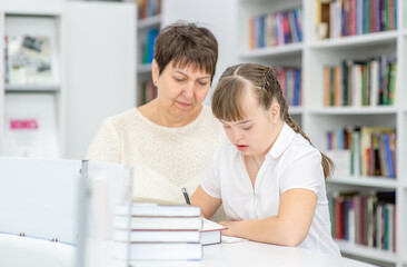 Fototapeta na wymiar Senior woman helps to girl with syndrome doing homework at a school. Education for disabled children concept