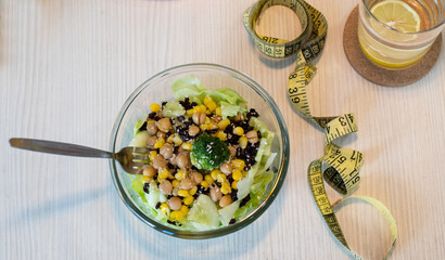 Fresh Salad in bowl and glass of water with lemon and measure tape. Chickpea with boiled corn, broccoli, lettuce, sesame. In the background is a plate with pumpkin seeds. Healthy lifestyle concept