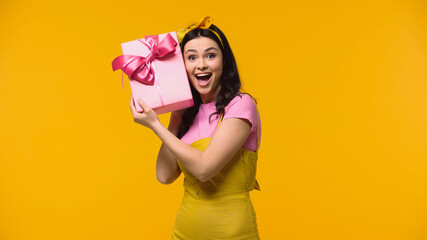 Astonished woman holding gift near ear isolated on yellow.