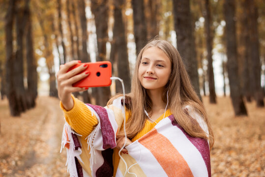 Happy blond girl taking selfie in an autumn park and listening to music. Teenage girl resting in the park alone, taking photos of herself and vibrant autumn nature around