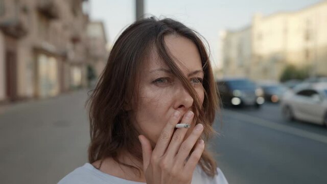 thoughtful woman with cigarette is standing near road in city, smoking and looking on car traffic