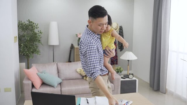 asian multitasking father working from home is coaxing his curious child in the arms while answering a phone call in the living room.
