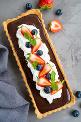 Chocolate cream tart with whipped cream, fresh blueberries and strawberries on top on a dark background. Summer dessert.