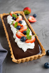 Chocolate cream tart with whipped cream, fresh blueberries and strawberries on top on a dark background. Summer dessert.