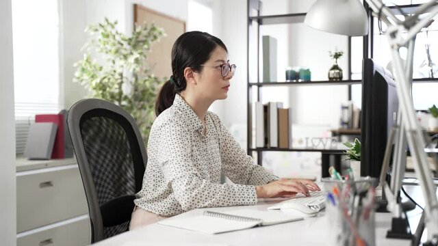 side portrait of an asian female manager working on the desktop computer with concentration at desk in a modern office with daylight.