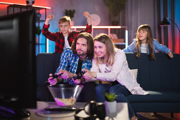 Excited husband and wife playing video games with joystick while son and daughter cheering them behind. Young caucasian family spending time at home with fun.
