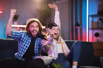 Cheerful young woman with man enjoying winning in video games while sitting on couch. Caucasian couple in casual outfit spending free time with fun at home.