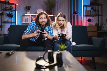 Competitive emotional smiling playful 30-aged Caucasian couple sitting on the blue sofa at home in the evening and playing videogames on TV using joysticks © sofiko14