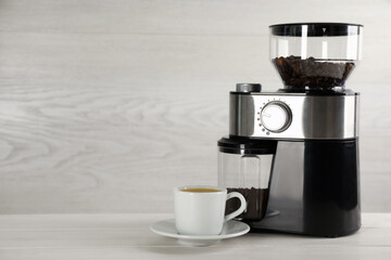 Modern electric coffee grinder and cup of fresh drink on white wooden table. Space for text