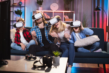 Family, leisure and technology concept. Funny smiling friendly family sitting on the sofa at home in the evening and using virtual reality headsets for video games or movie