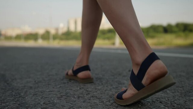 woman is walking on asphalt road in city, closeup of female feet in sandals