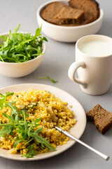Delicious bulgur porridge with fried onions and herbs in a plate, a glass of milk, arugula in a bowl and black bread