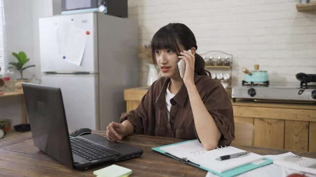 Korean female receiving notification on the phone is cheering with clenched fists for winning business opportunity while working from home on the laptop at table