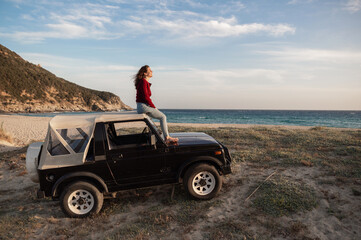 Young woman sitting on rooftop of her off-road car enjoying the wind and looking at the sea. Car parked in front of a wild beach.
