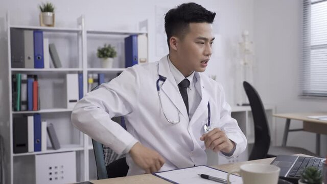 slow movement Taiwanese physician suffering lumbago is looking into distance while exercising hid stiff body and massaging lower back at desk in clinic office