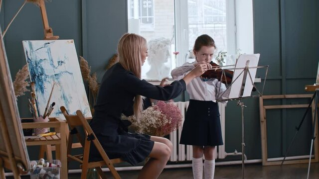Young blonde woman teacher teaching a little girl how to properly hold a bow while playing violin - class lesson