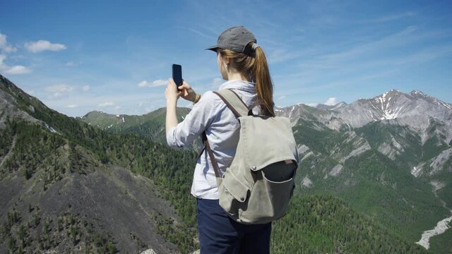 Woman traveler in a cap with a hiking backpack takes pictures of the mountains using the phone. She is saddened by the incredible beauty of the high mountains. Panoramic shot. 4K UHD