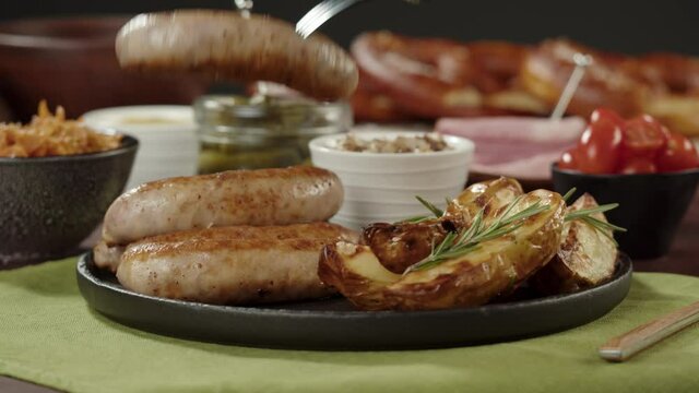 Taking smoked sausages with fork close-up, fried potatoes, pickled cucumbers, bratwursts and fresh pretzels on table. Traditional German Cuisine. Composition of Cooked National Czech Food. 