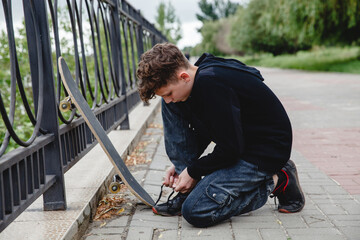 A curly-haired European teen in a black hoodie with skate sitting on the embankment and tying his shoelaces on sneakers  - obrazy, fototapety, plakaty