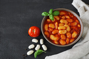 large white beans cooked in tomatoes and tomato paste in a brown bowl with basil leaves on dark background with linen napkin