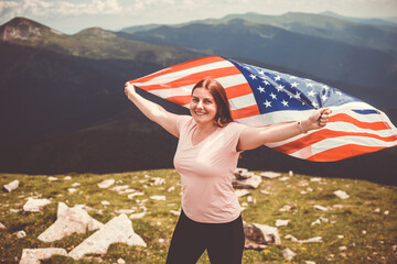 Girl with American Flag looking out at landscape. Young woman stands in the mountains, holding the US flag in her arms high. Flag fluttering in the wind. Toned image