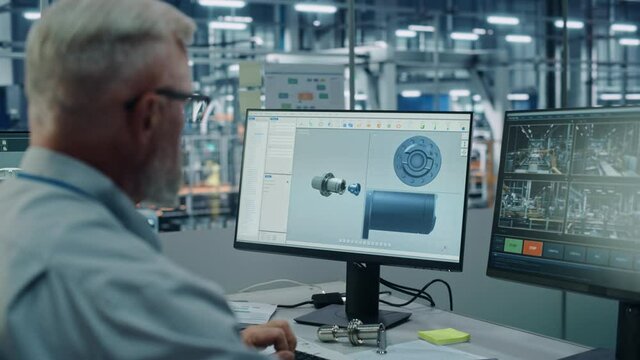 Car Factory Office: Male Chief Automotive Engineer Sitting at His Desk Working on Computer. Monitoring, Control, Equipment Design. Automated Robot Arm Assembly Line Manufacturing Vehicles