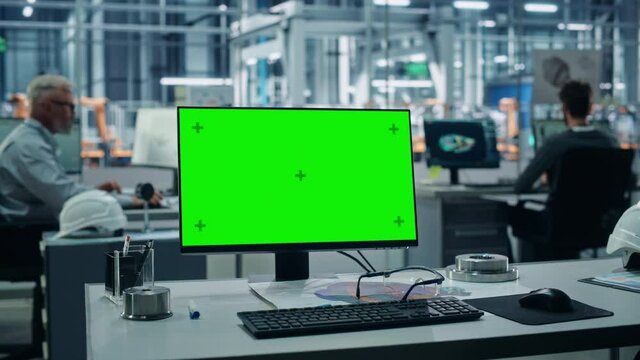 Car Factory: On the Desk Green Screen Chroma Key Computer. In Background Diverse Team of Engineers Work in Office of Automated Robot Arm Assembly Line Manufacturing Vehicles