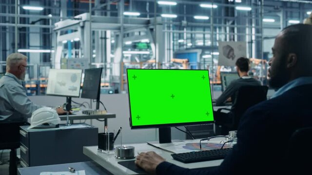 Car Factory Office: Black Male Chief Automotive Engineer Sitting at His Desk Working on Green Screen Chroma Key Computer. Automated Robot Arm Assembly Line Manufacturing. Over Shoulder Medium Shot