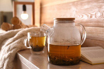 Glass teapot and cup of hot tea near open book on wooden table in room. Cozy home atmosphere