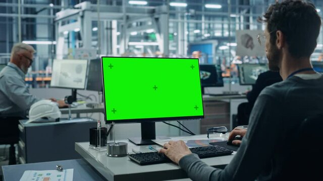 Car Factory Office: Caucasian Male Automotive Engineer Sitting at His Desk Working on Green Screen Chroma Key Computer. Automated Robot Arm Assembly Line Manufacturing. Over Shoulder Medium Shot