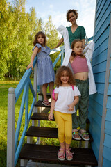 Fototapeta na wymiar Cheerful woman with adorable girls standing on stairs outside blue cottage on sunny summer day in countryside