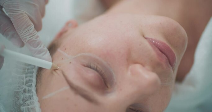 A cosmetologist is using a cosmetic pencil for painting a white line on the face of a female client before the procedure.
