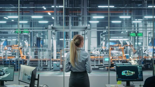 Car Factory Office: Successful Female Chief Engineer Overlooking Automated Robot Arm Assembly Line Manufacturing Advanced High-Tech Electric Vehicles. Production Conveyor. Back View Shot