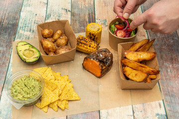 Chef's hands placing onion on a salad in a set of Mexican-style takeout plates with avocado, tortilla chips, guacamole, roasted sweet potato, grilled corn, and fried and roasted potatoes