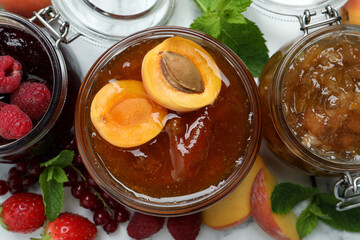 Jars with different jams and fresh fruits on white table, flat lay