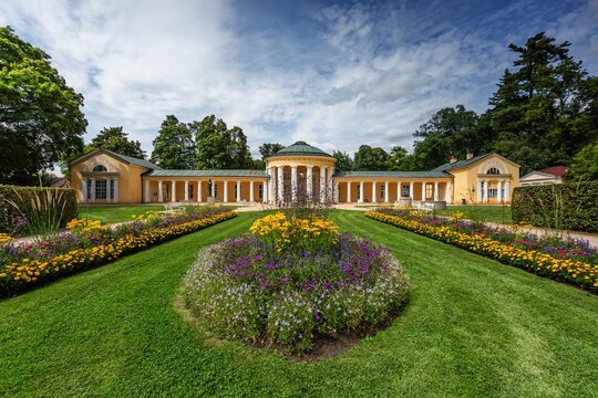 Marienbad, Czech Republic - August 12 2021: View of the yellow Ferdinand spring pavilion with columns, colorful flowers and mowed green lawn. Summer sunny day in spa city. Blue sky with white clouds.