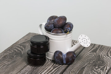 Large plums in a metal watering can. On pine boards. Nearby is a jar of jam. Harvest plums.