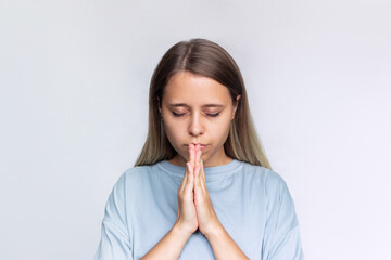 A young caucasian blonde woman in a gray t-shirt prays with her eyes closed and folded hands, thanks, making wish, asking help, hope or forgiveness isolated on a white background