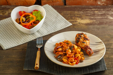 Seashell pasta in a tomato with a grilled chicken leg on a stand and a napkin next to a fork and a vegetable salad.