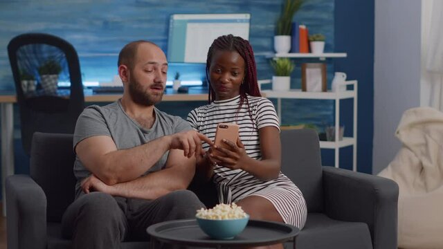 Interracial couple looking at smartphone screen at home. Young multi ethnic partners using modern device with technology while sitting on living room couch. Mixed race people with gadget