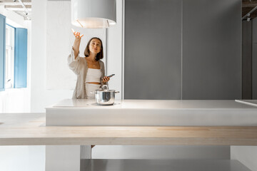 Young woman controls hood, while standing with cell phone and cooking at modern kitchen. Concept of...
